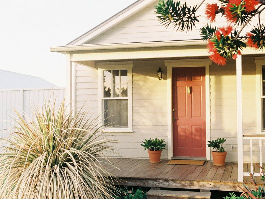 A wooden front porch with a red door, potted plants and a pohutukawa branch